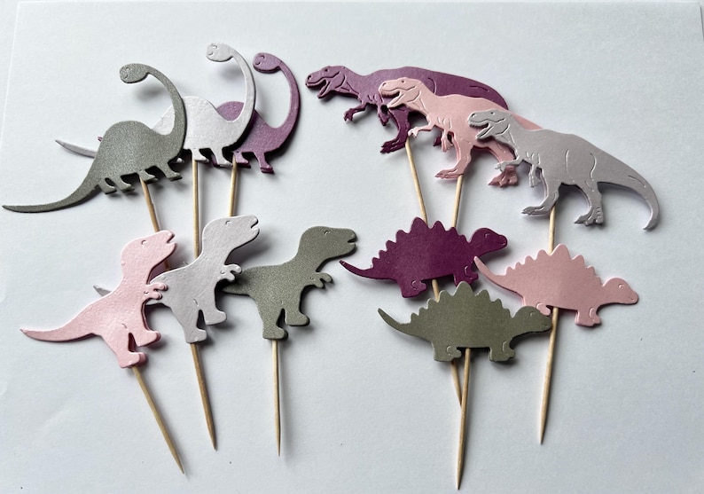 May include: A set of 10 dinosaur cupcake toppers in pink, purple, and gray. The toppers are made of cardstock and are attached to wooden skewers.