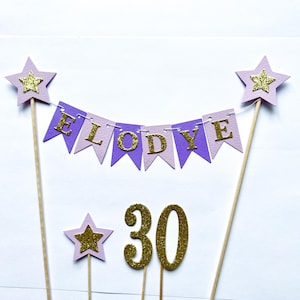 May include: A cake topper set featuring a bunting banner with purple and light purple pennants spelling ELODYE in gold glitter letters, two large stars on wooden skewers, a small star on a skewer, and the number 30 in gold glitter.