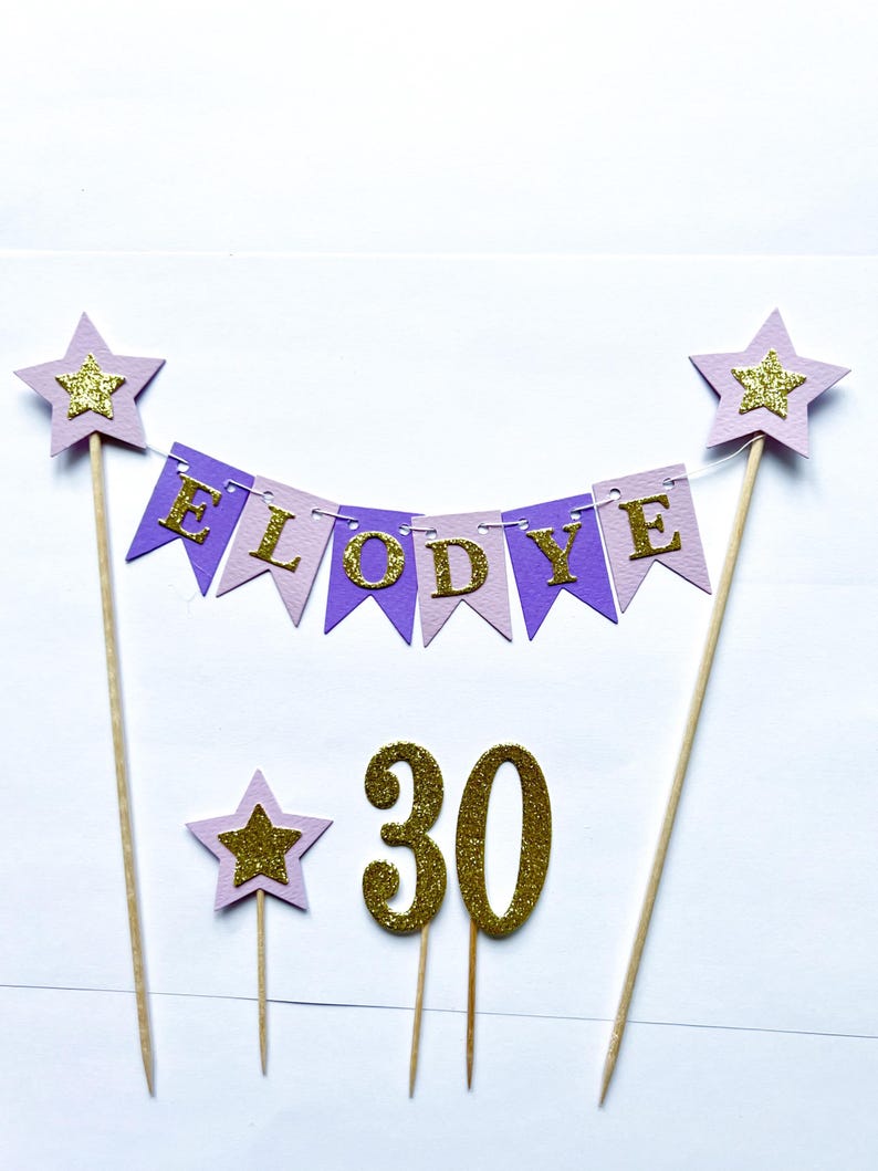 May include: A cake topper set featuring a bunting banner with purple and light purple pennants spelling ELODYE in gold glitter letters, two large stars on wooden skewers, a small star on a skewer, and the number 30 in gold glitter.
