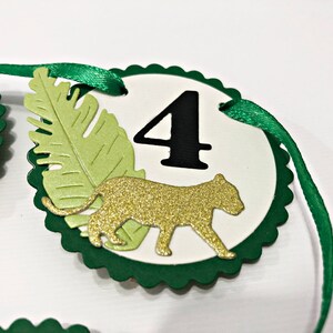 May include: Green and white circle with a scalloped edge, featuring a gold glitter cheetah silhouette, a green leaf, and the number "4" in black. The circle is attached to a green ribbon.
