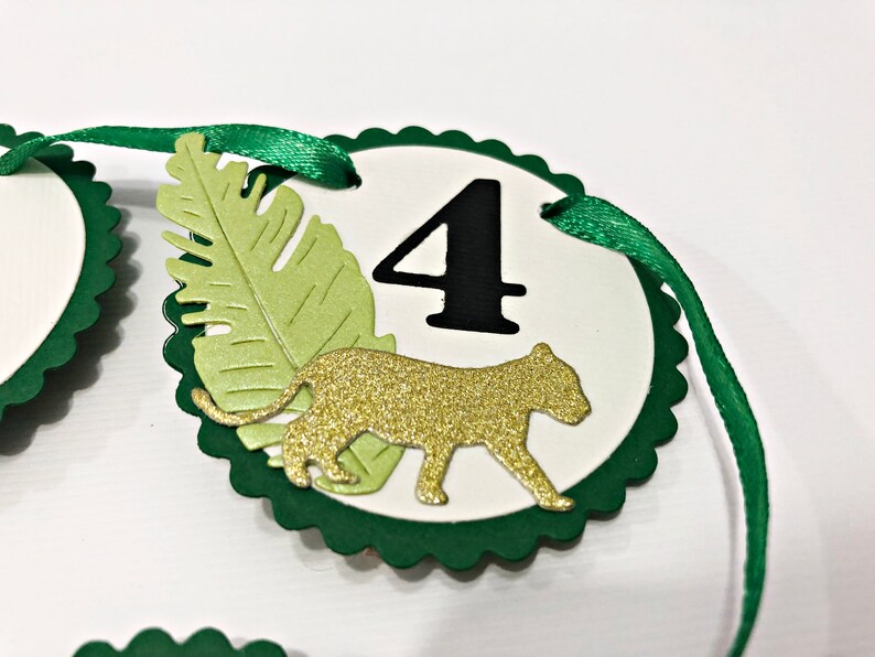 May include: Green and white circle with a scalloped edge, featuring a gold glitter cheetah silhouette, a green leaf, and the number "4" in black. The circle is attached to a green ribbon.