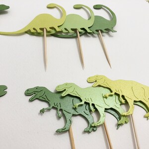 May include: Green and yellow dinosaur cupcake toppers. The toppers are made of paper and have a glossy finish. They are attached to wooden skewers.