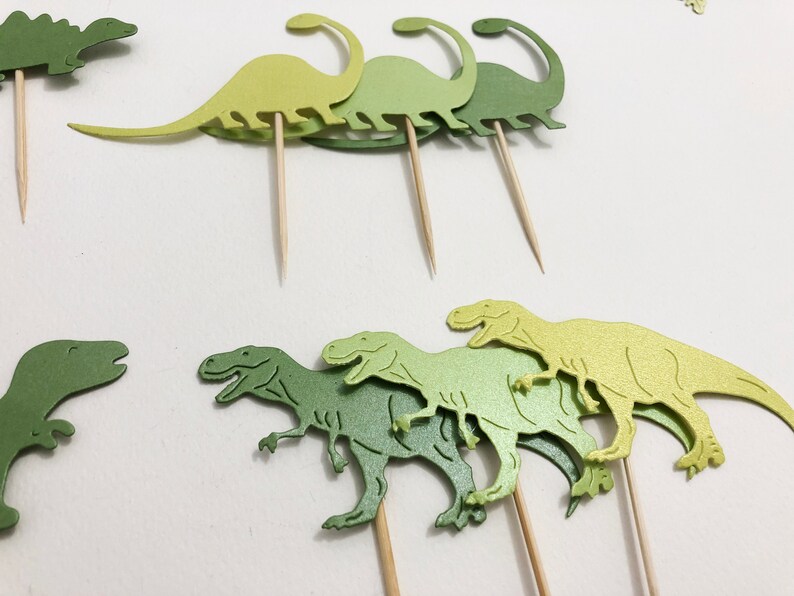 May include: Green and yellow dinosaur cupcake toppers. The toppers are made of paper and have a glossy finish. They are attached to wooden skewers.