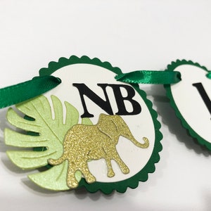 May include: Green and white banner with the letters "NB" and a gold glitter elephant. The banner is decorated with green leaves.