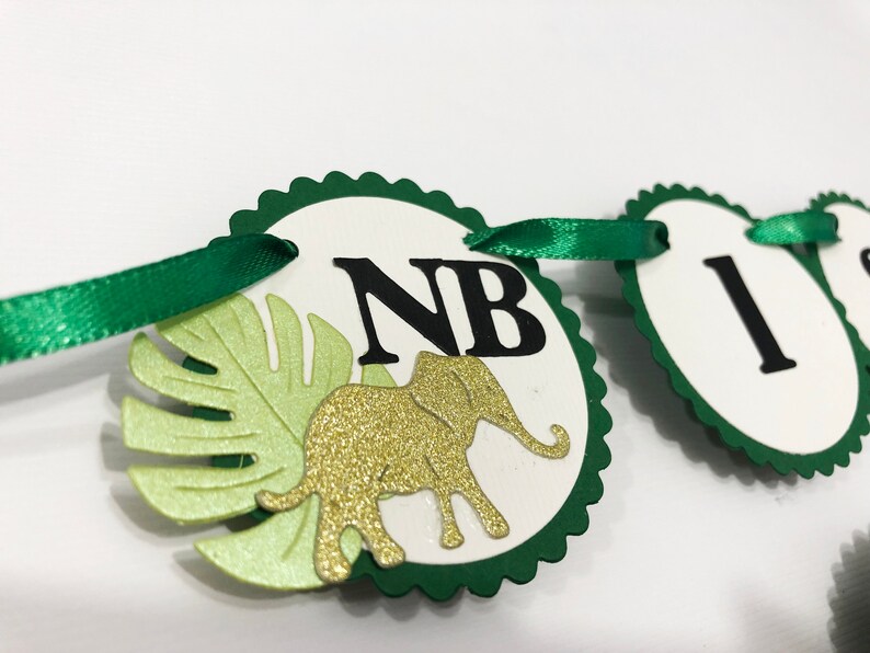 May include: Green and white banner with the letters "NB" and a gold glitter elephant. The banner is decorated with green leaves.