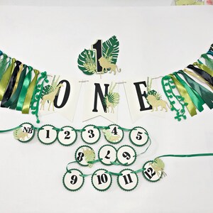 May include: Green and gold jungle-themed first birthday party decorations. The banner says "ONE" with a lion, giraffe, and palm leaves. There are also 12 round green and white circles with numbers 1 through 12 and a "NB" for newborn.