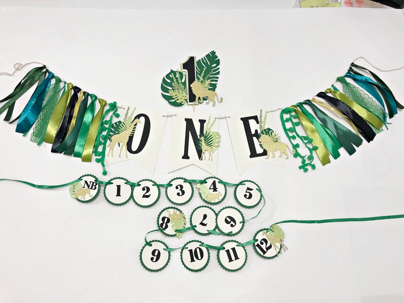 May include: Green and gold jungle-themed first birthday party decorations. The banner says "ONE" with a lion, giraffe, and palm leaves. There are also 12 round green and white circles with numbers 1 through 12 and a "NB" for newborn.