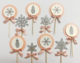 May include: A collection of cupcake toppers with a winter theme. The toppers feature a light peach scalloped circle with a white center, adorned with a silver glitter snowflake or a baby bottle design. Each topper has a peach satin bow.