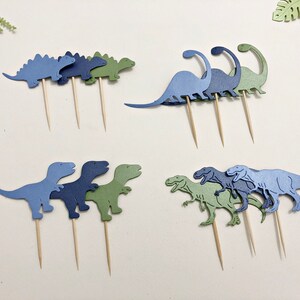 May include: A set of nine dinosaur cupcake toppers in blue and green. The toppers are made of paper and are attached to wooden skewers. The dinosaurs are in various poses, including standing, walking, and running.