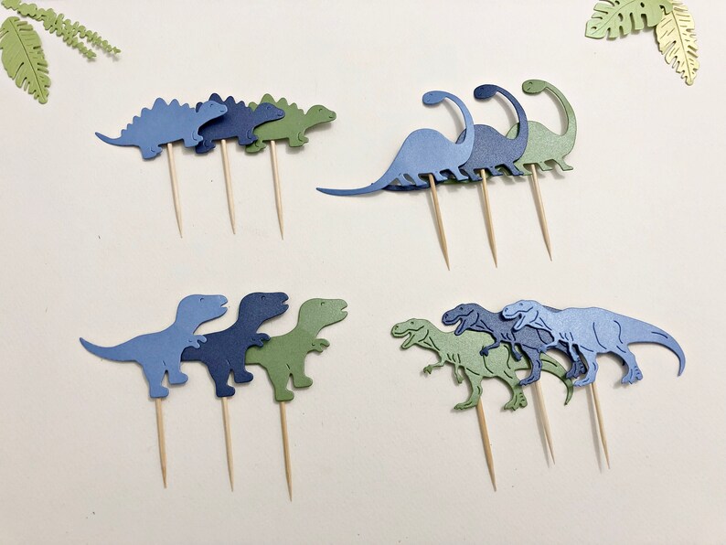 May include: A set of nine dinosaur cupcake toppers in blue and green. The toppers are made of paper and are attached to wooden skewers. The dinosaurs are in various poses, including standing, walking, and running.