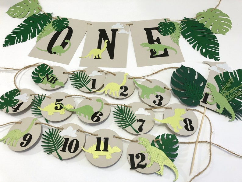 K&ouml;nnte beinhalten: Ein gr&uuml;nes und braunes Dinosaurier-Motto-Banner f&uuml;r die erste Geburtstagsfeier mit dem Wort "ONE" in schwarzen Buchstaben. Das Banner ist mit gr&uuml;nen Bl&auml;ttern und Dinosauriern bespannt. Es gibt auch 12 runde Papierkreise mit den Zahlen 1 bis 12, gr&uuml;nen Bl&auml;ttern und Dinosauriern.