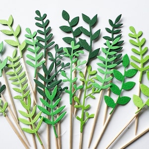 Può includere: Un set di foglie di carta verde su spiedini di legno. Le foglie sono di varie tonalità di verde e sono disposte a forma di bouquet.