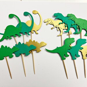 May include: A set of 10 green and gold dinosaur cupcake toppers. The dinosaurs are in various poses, including standing, walking, and running. The toppers are perfect for a dinosaur-themed party.