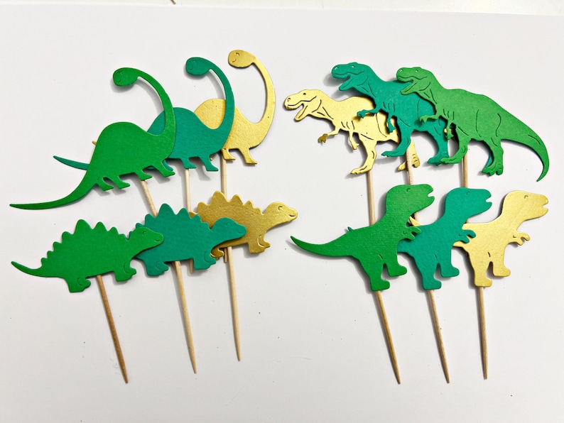 May include: A set of 10 green and gold dinosaur cupcake toppers. The dinosaurs are in various poses, including standing, walking, and running. The toppers are perfect for a dinosaur-themed party.