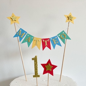 A cake topper featuring a bunting banner with the name MATTEO in gold glitter letters on colorful pennants, suspended between two wooden sticks topped with yellow stars, plus a gold glitter number 1 and a red star on sticks.