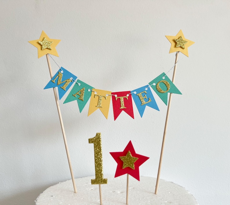 A cake topper featuring a bunting banner with the name MATTEO in gold glitter letters on colorful pennants, suspended between two wooden sticks topped with yellow stars, plus a gold glitter number 1 and a red star on sticks.