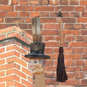 Könnte beinhalten: Eine skurrile Holzskulptur einer Person, die einen schwarzen Zylinder und einen braunen Mantel trägt. Die Skulptur steht vor einer Backsteinmauer.