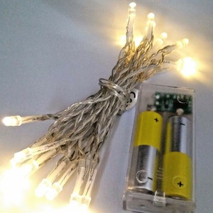 Könnte beinhalten: Eine Lichterkette mit warmweißen LED-Lichtern und einem Batteriefach. Die Lichter sind um das Batteriefach gewickelt und einsatzbereit.