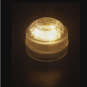 Könnte beinhalten: Ein kreisförmiger, klarer LED-Licht-Puck aus Kunststoff mit gewölbter Oberseite und geripptem Seitenrand. Das Gerät strahlt von innen ein warmes, helles gelbes Licht aus, das die durchscheinende Oberfläche beleuchtet und sanftes Licht abgibt.