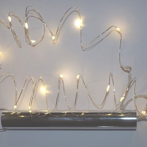 Könnte beinhalten: Eine Lichterkette mit kleinen, warmweißen LED-Leuchten an einem dünnen, silbernen Draht. Die Lichter werden von einem silbernen, zylindrischen Batteriepack mit gebogenem Griff gespeist. Die Lichter sind in einem wellenförmigen Muster angeordnet.