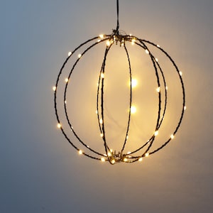 Könnte beinhalten: Eine hängende dekorative Kugel mit warmweißen LED-Leuchten. Die Kugel besteht aus dunklen Metallringen, die ein käfigartiges Design bilden. Die Lichter sind gleichmäßig verteilt und erzeugen ein sanftes Leuchten. Ideal für die Inneneinrichtung oder Festbeleuchtung.