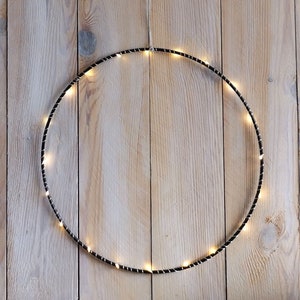 Könnte beinhalten: Ein schwarzer Drahtkreis mit kleinen weißen Lichtern am Rand, der vor einem hellen Holzhintergrund hängt.