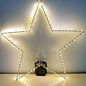 K&ouml;nnte beinhalten: Eine sternf&ouml;rmige Leuchte mit warmwei&szlig;en LED-Lichtern. Der Stern ist mit einem dunklen Draht umrandet und hat unten einen kleinen schwarzen Akku. Der Stern h&auml;ngt vor einer wei&szlig;en Wand. Die Abmessungen des Sterns betragen ca. 60 cm.