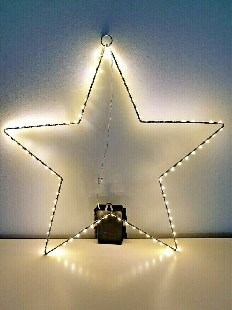 K&ouml;nnte beinhalten: Eine sternf&ouml;rmige Leuchte mit warmwei&szlig;en LED-Lichtern. Der Stern ist mit einem dunklen Draht umrandet und hat unten einen kleinen schwarzen Akku. Der Stern h&auml;ngt vor einer wei&szlig;en Wand. Die Abmessungen des Sterns betragen ca. 60 cm.