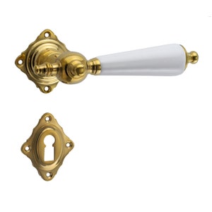 May include: A gold door handle with a white ceramic lever. The handle has a decorative, diamond-shaped backplate with a keyhole cover.