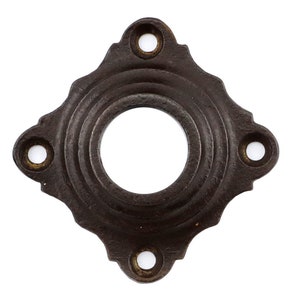 Op de afbeelding: Een donkerbruine metalen deurknop rozet met een vierkante vorm en afgeronde hoeken. De rozet heeft een decoratief, concentrisch cirkelontwerp en vier kleine gaten rond de omtrek.