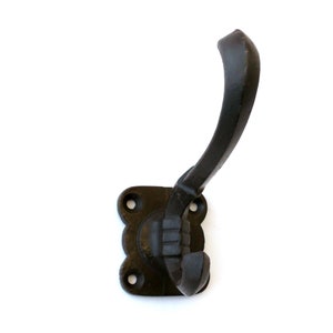 Könnte beinhalten: Schwarzer Haken aus Gusseisen mit quadratischem Sockel und gebogenem Haken. Der Haken ist mit einem dekorativen Design an der Basis befestigt.