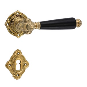 Puede incluir: Una manilla de puerta dorada con una palanca de madera negra y una tapa de mirilla a juego. La manilla tiene un diseño ornamentado con detalles intrincados.