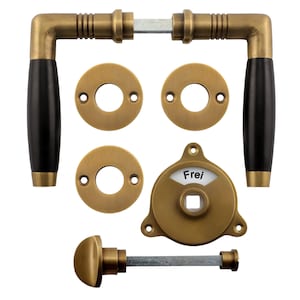 May include: A set of antique brass door hardware with black wooden handles. The set includes two door handles, two round escutcheons, a round escutcheon with a keyhole, and a round knob with a lever. The escutcheons have three small holes each. The keyhole escutcheon has the word "Frei" written on it.