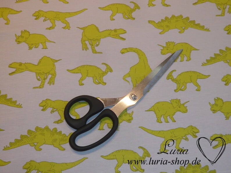 May include: A white fabric with a repeating pattern of yellow dinosaurs. A pair of silver scissors is lying on top of the fabric. The text "Luria www.luria-shop.de" is printed on the fabric.