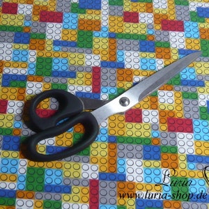 Puede incluir: Un par de tijeras con mangos negros sobre un fondo de tela colorido con un patr&oacute;n repetitivo de cuadrados y c&iacute;rculos. Los cuadrados son de varios colores, incluyendo rojo, amarillo, azul, verde y gris. Los c&iacute;rculos son blancos.