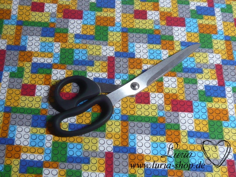 Puede incluir: Un par de tijeras con mangos negros sobre un fondo de tela colorido con un patr&oacute;n repetitivo de cuadrados y c&iacute;rculos. Los cuadrados son de varios colores, incluyendo rojo, amarillo, azul, verde y gris. Los c&iacute;rculos son blancos.