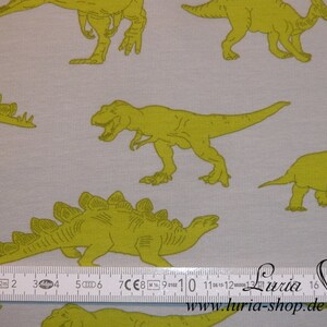 May include: A white fabric with a repeating pattern of green dinosaurs. The dinosaurs are in various poses, including walking, standing, and roaring. A ruler is included in the image for scale.