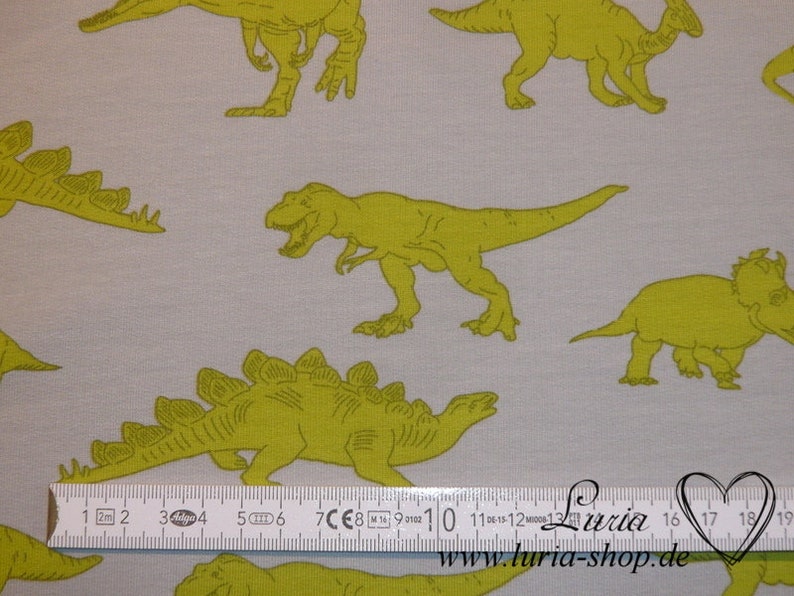 May include: A white fabric with a repeating pattern of green dinosaurs. The dinosaurs are in various poses, including walking, standing, and roaring. A ruler is included in the image for scale.
