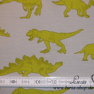 May include: A white fabric with a repeating pattern of green dinosaurs. The dinosaurs are in various poses, including walking, standing, and roaring. A ruler is included in the image for scale.