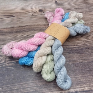 Könnte beinhalten: Ein Set mit fünf Garnknäuel in den Farben Rosa, Blau, Grün und Beige. Das Garn ist um ein braunes Papier-Etikett mit dem aufgedruckten Text "Scents & Threads" und "Hand-dyed Yarn" gewickelt.