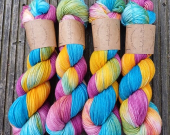 May include: Four skeins of hand-dyed yarn in a rainbow colorway. The yarn is a mix of yellow, orange, pink, blue, and green. The skeins are labeled "Liebe Handarbeit" and "hubbystreet".