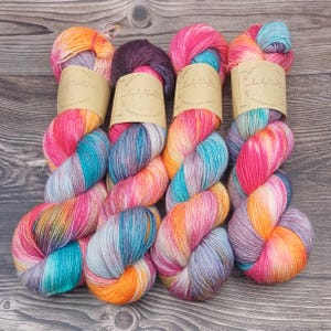 Könnte beinhalten: Vier Garnstränge in handgefärbter, lebendiger Farbpalette aus leuchtendem Pink, Orange, Türkis und zartem Violett. Jeder Strang ist mit einer beigen Papierbanderole versehen, die mit dem Text "bridgetwalt" und "schönes aus wolle" bedruckt ist.