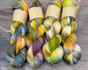May include: Four skeins of hand-dyed yarn arranged side by side. Each skein features a variegated color palette of yellow, green, purple, and grey tones. Every skein is secured with a tan paper label featuring printed text and a logo.