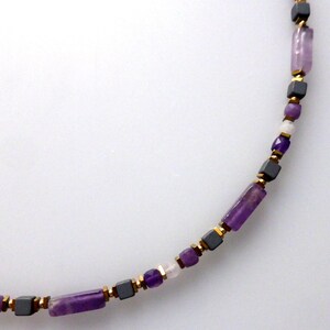 Könnte beinhalten: Eine Halskette mit einer Mischung aus lila, grauen und goldenen Perlen. Die Halskette hat rechteckige und würfelförmige Perlen sowie kleinere runde und facettierte Perlen. Die Perlen sind mit goldfarbenen Metallakzenten aufgefädelt.