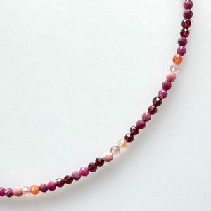 Könnte beinhalten: Eine zarte Halskette aus kleinen, facettierten Perlen in Rosa-, Rot- und Orangetönen. Die Perlen sind aufgereiht und erzeugen einen subtilen Farbverlauf. Die Halskette wird vor einem weißen Hintergrund präsentiert.