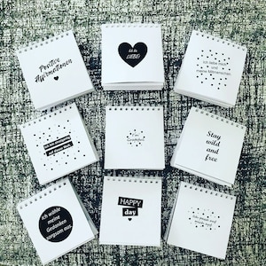 Könnte beinhalten: Neun kleine weiße Notizbücher mit schwarzer Spiralbindung. Jedes Notizbuch hat einen anderen inspirierenden Spruch oder Satz auf der Vorderseite gedruckt. Die Sprüche beinhalten "Positive Affirmationen", "Ich bin dankbar", "Bleib wild und frei", "Glücklicher Tag", "Ich wähle meine Gedanken sorgfältig aus", "Ich liebe mich selbst und meine Mitmenschen", "Ich bin einzigartig auf dieser Welt", und "Ich liebe mich selbst".