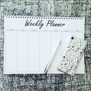 Könnte beinhalten: Ein weißer Wochenplaner mit dem schwarzen Titel "Weekly Planner" und den Wochentagen, die obenauf beschriftet sind. Ein silberner Stift und eine silberne Handyhülle mit geometrischem Muster liegen auf dem Planer.
