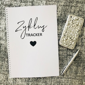 Könnte beinhalten: Ein weißes Spiralnotizbuch mit dem Text "Zyklus TRACKER" und einem schwarzen Herzen. Das Notizbuch liegt auf einer grauen strukturierten Oberfläche mit einer silbernen Handyhülle und einem silbernen Stift.