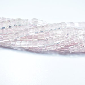 Puede incluir: Primer plano de un hilo de cuentas cuadradas de color rosa claro. Las cuentas son translúcidas y parecen estar hechas de vidrio o un material similar. Las cuentas están ensartadas, creando un patrón fluido y texturizado.
