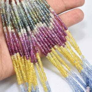 Può includere: Fili di perline di pietre preziose sfaccettate in una varietà di colori, tra cui giallo, viola, blu e verde. Le piccole perline sono infilate insieme, creando un'esposizione strutturata e colorata. Probabilmente zaffiri.